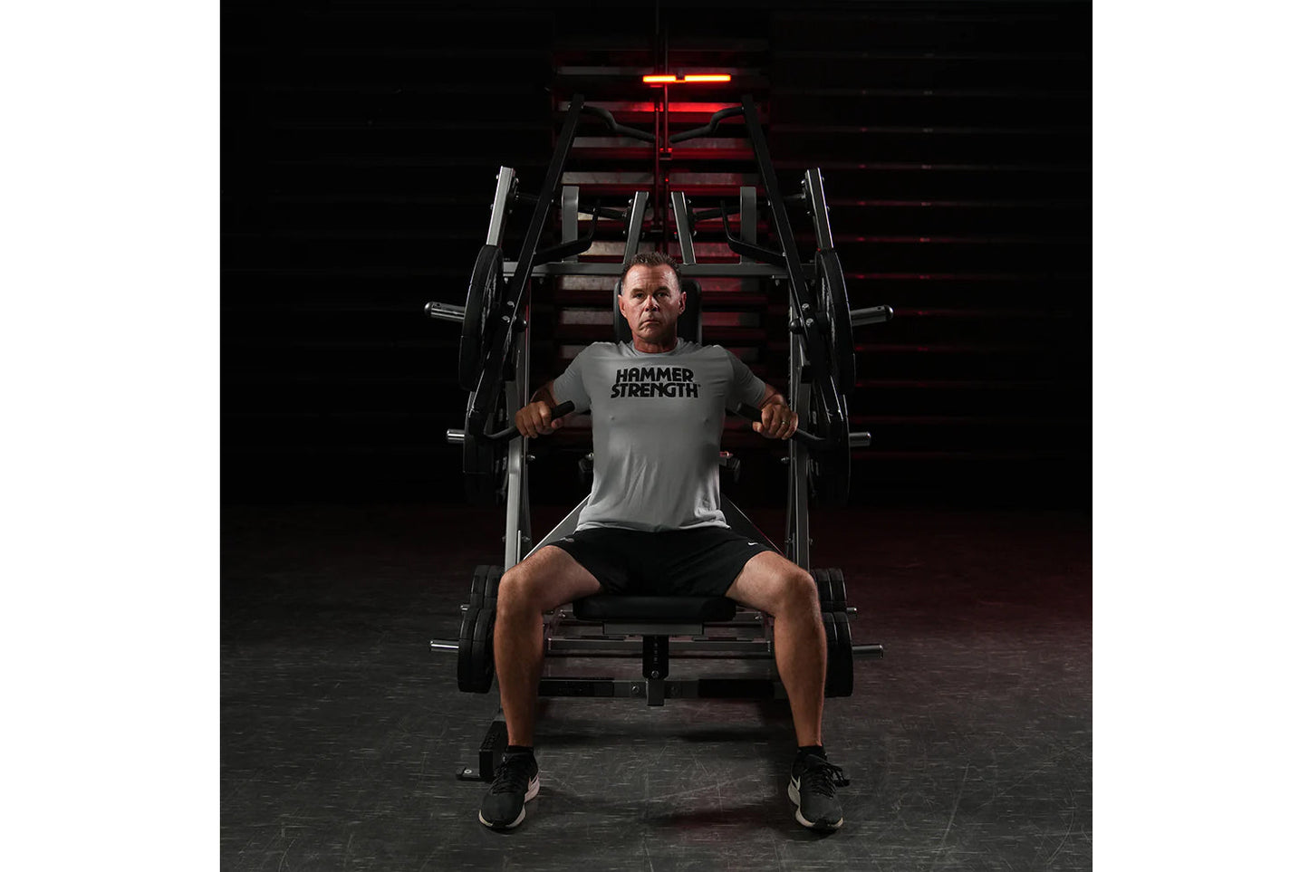Man wearing gray Hammer Strength t-shirt and black shorts using plate-loaded chest machine in gym with red lighting