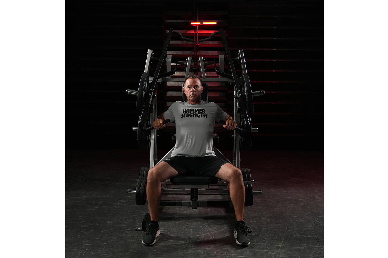 Man wearing gray Hammer Strength t-shirt and black shorts using plate-loaded chest machine in gym with red lighting