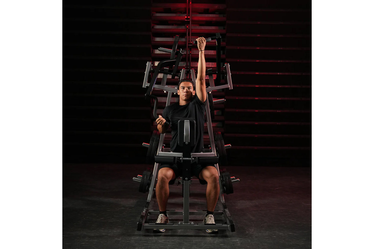 Man in black shirt and shorts performing lat pulldown exercise on Hammer Strength plate-loaded machine with red-lit backgroun