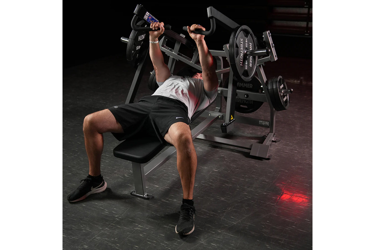 Man performing chest press exercise on black Hammer Strength plate-loaded bench with loaded barbell in gym setting