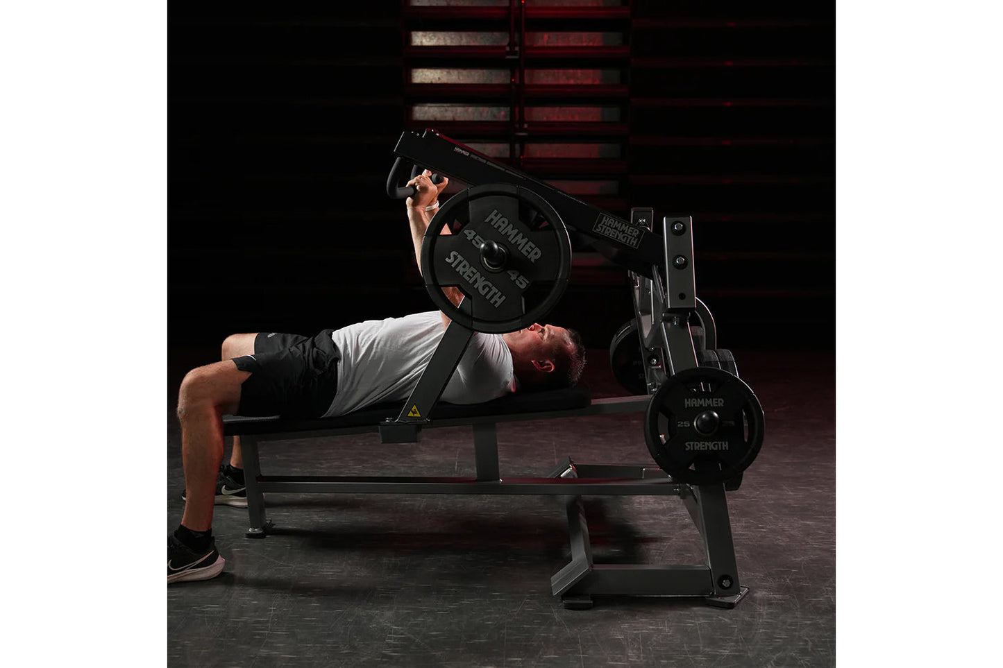 Male athlete performing chest press exercise on black Hammer Strength plate-loaded bench with weights loaded, in gym setting