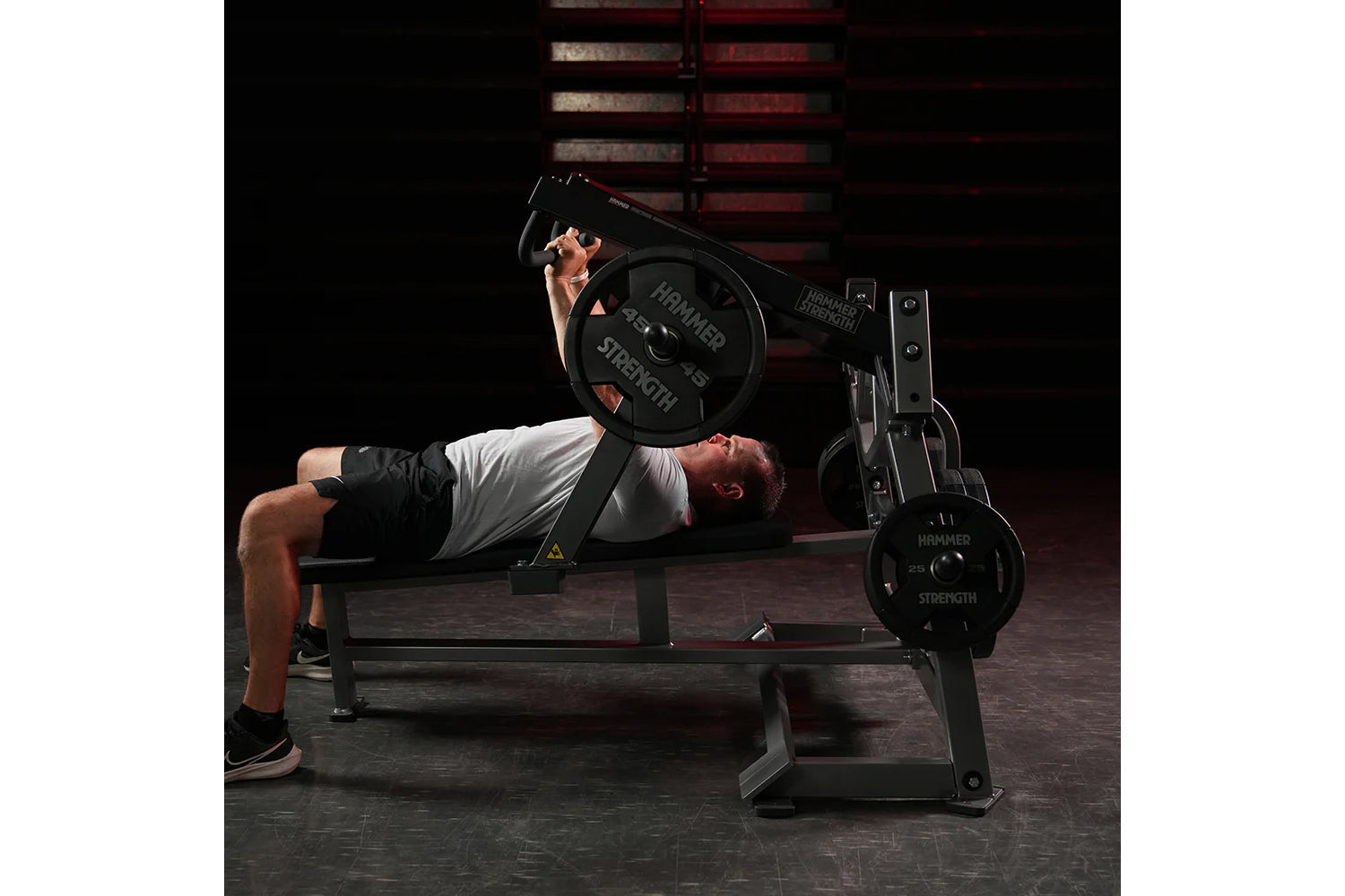 Male athlete performing chest press exercise on black Hammer Strength plate-loaded bench with weights loaded, in gym setting