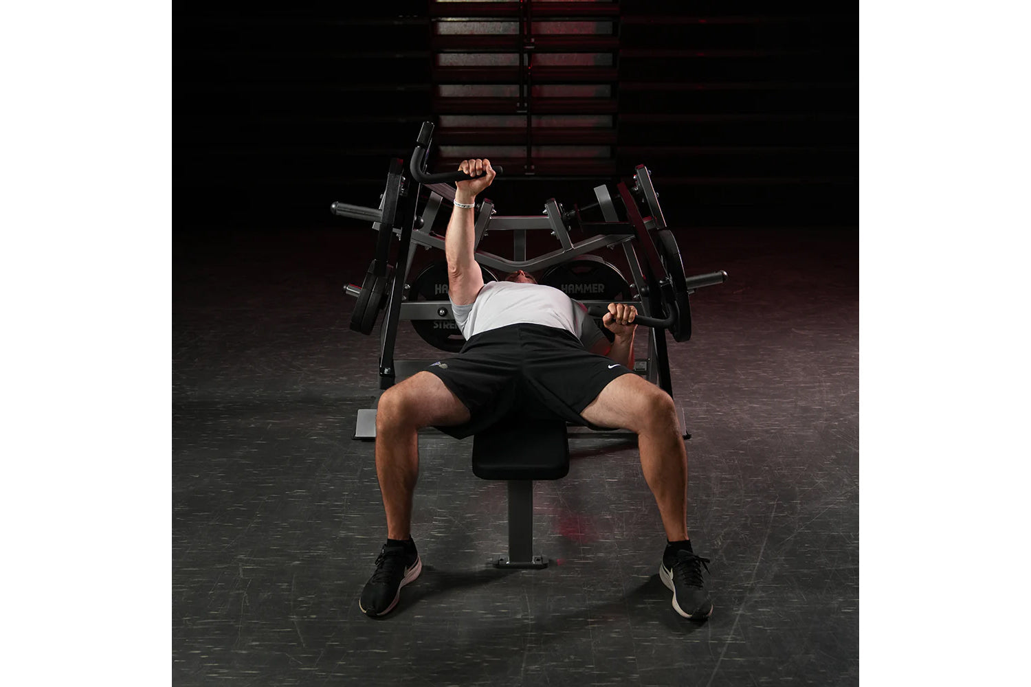 Man using black Hammer Strength plate-loaded chest press machine in gym, wearing white tank top and black shorts