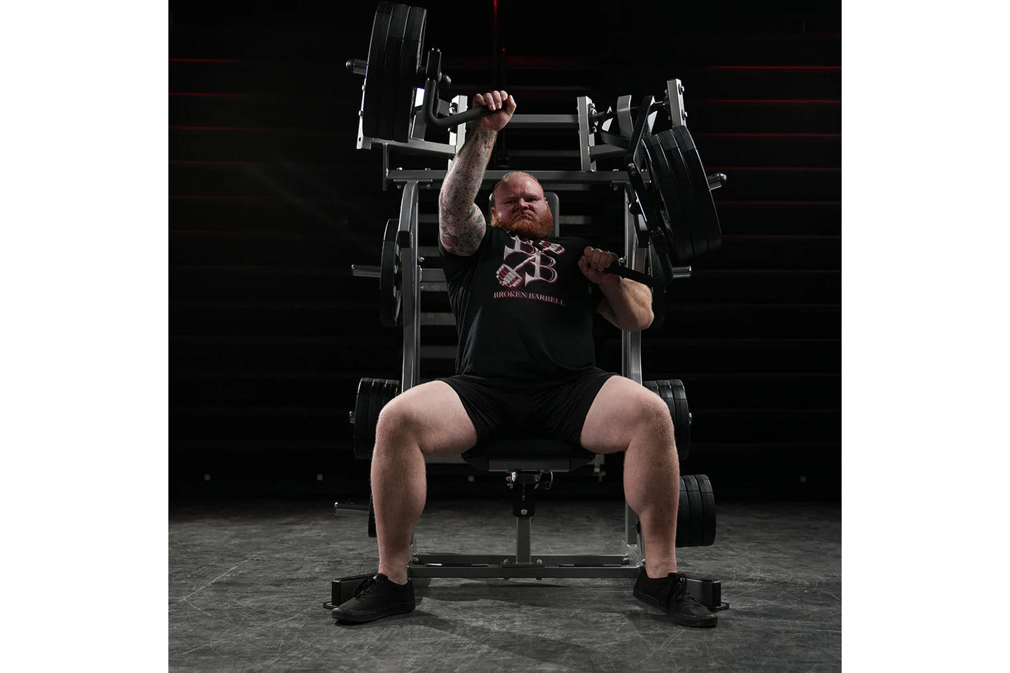 Man wearing black Broken Barrel t-shirt performing incline press exercise on plate-loaded weight machine in gym setting