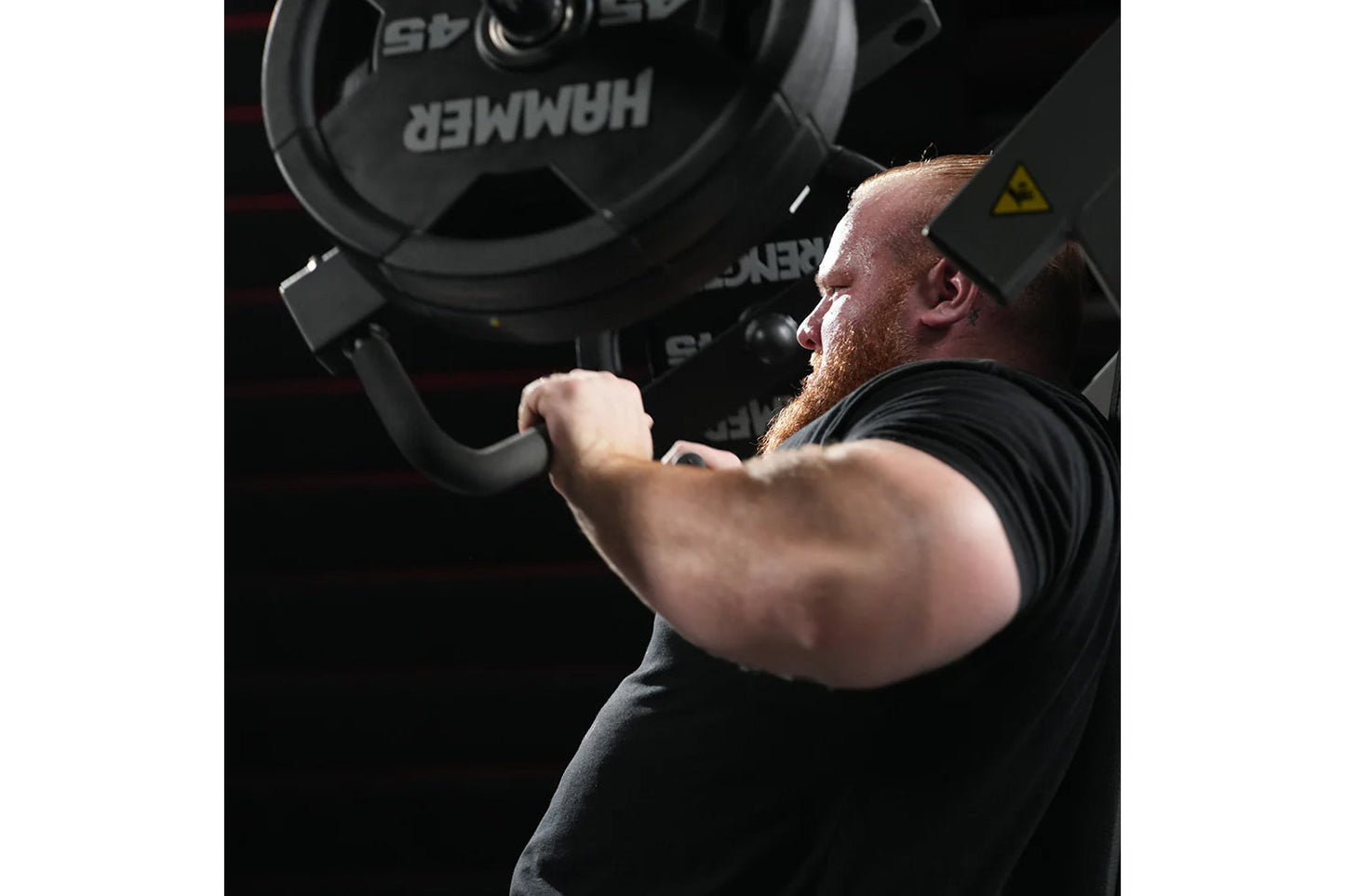 Man with red beard wearing black cap and shirt lifting heavy black dumbbell in gym setting