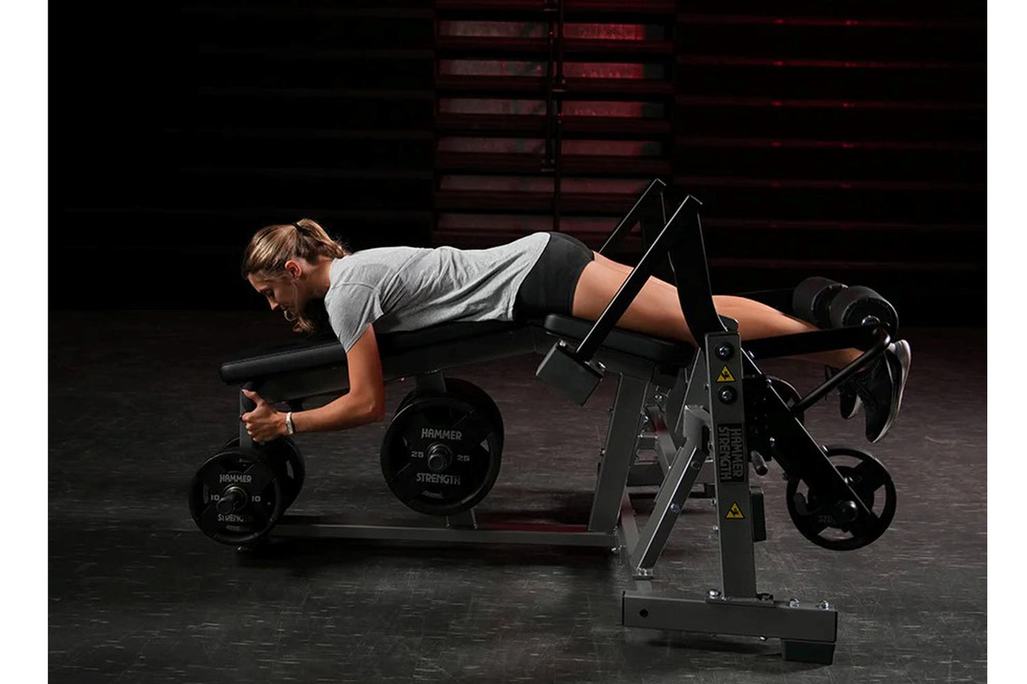 Woman using black plate-loaded leg curl machine with adjustable foot platform and weight plates, wearing gray t-shirt and bla