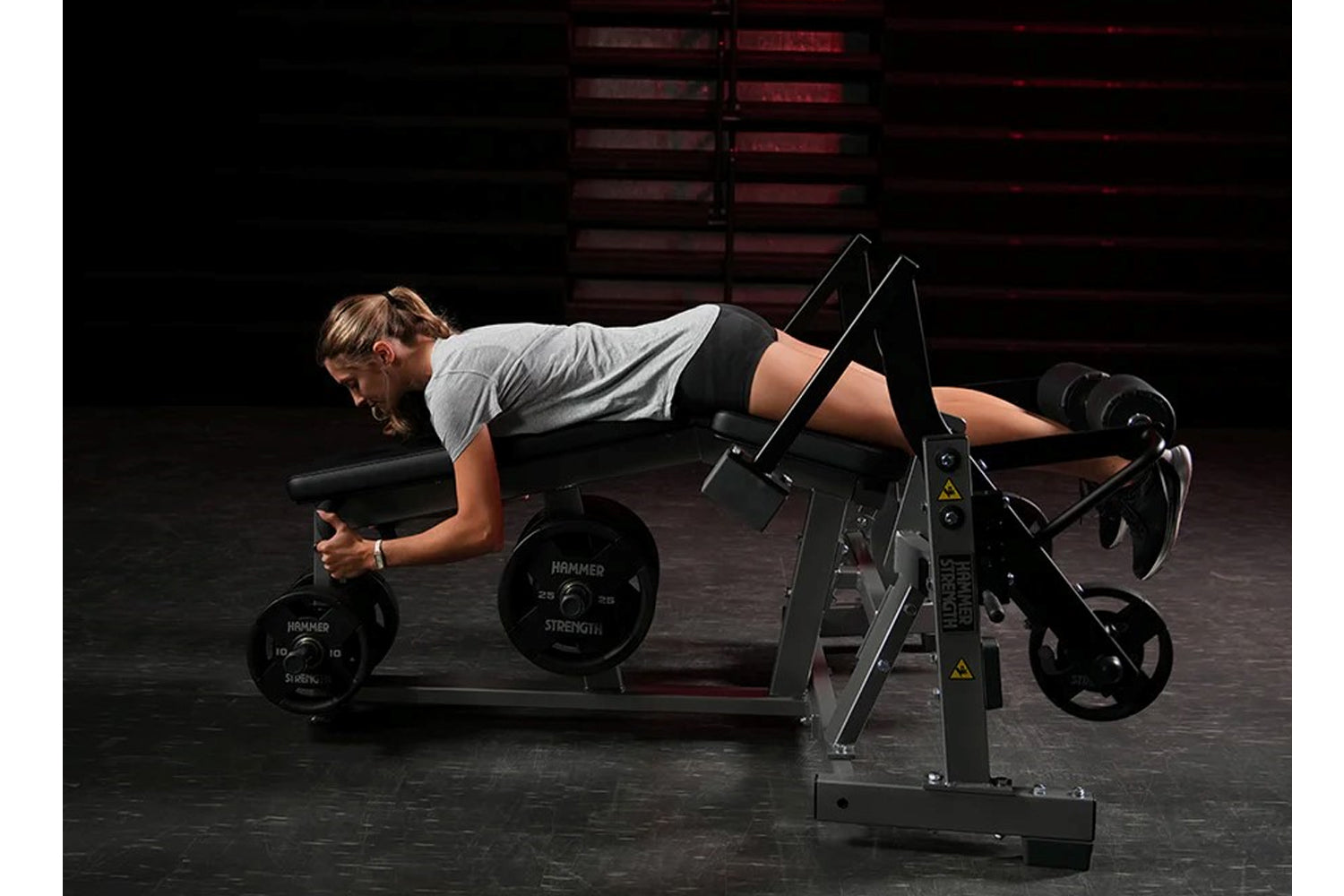 Woman using black plate-loaded leg curl machine with adjustable foot platform and weight plates, wearing gray t-shirt and bla
