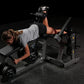 Woman performing leg curl exercise on black Hammer Strength plate-loaded machine while holding dumbbell, wearing gray shirt a