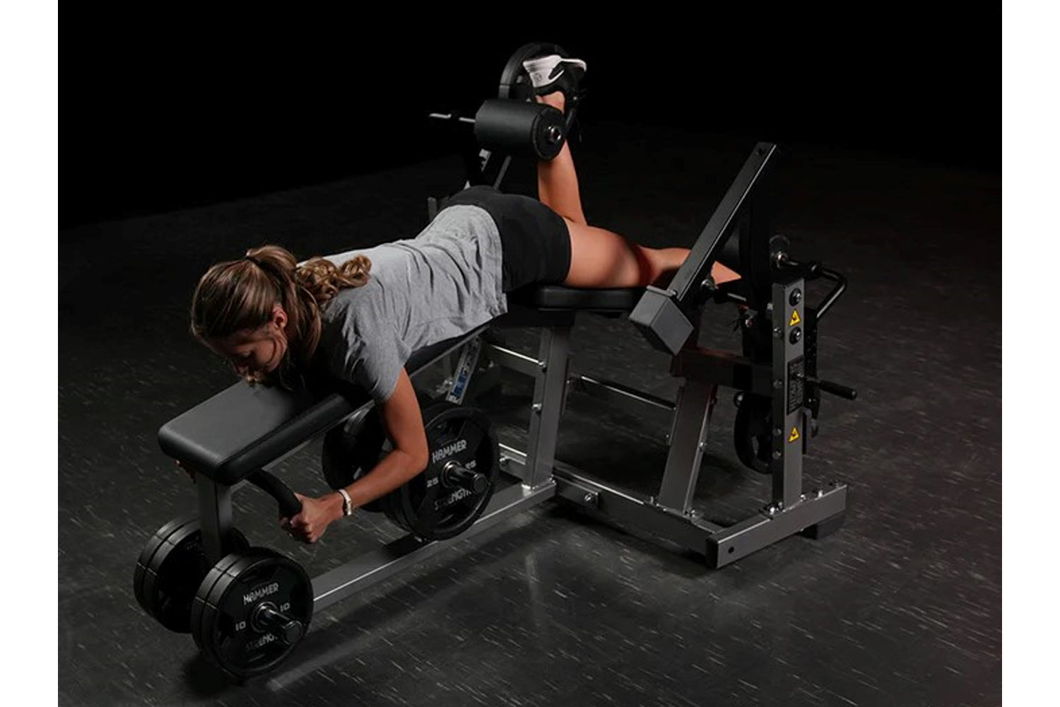 Woman performing leg curl exercise on black Hammer Strength plate-loaded machine while holding dumbbell, wearing gray shirt a