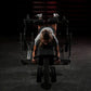 Person using black Hammer Strength plate-loaded leg curl machine in gym setting with dumbbells visible in background