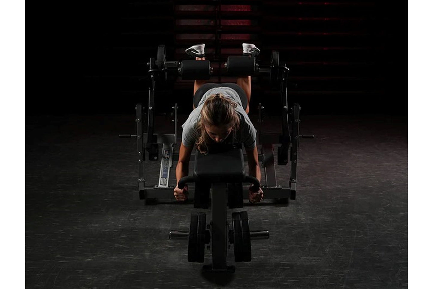 Person using black Hammer Strength plate-loaded leg curl machine in gym setting with dumbbells visible in background