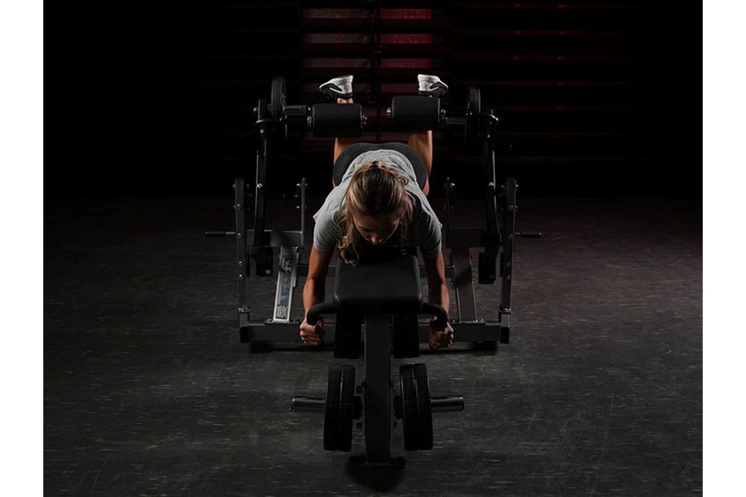 Person using black Hammer Strength plate-loaded leg curl machine in gym setting with dumbbells visible in background