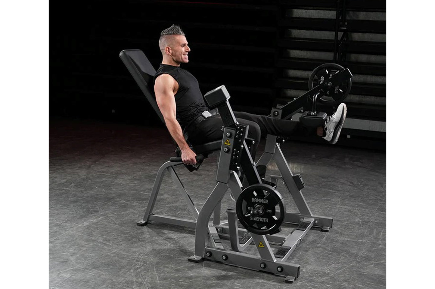 Man in black sleeveless shirt using Hammer Strength plate-loaded leg extension machine with weighted plates in gym setting
