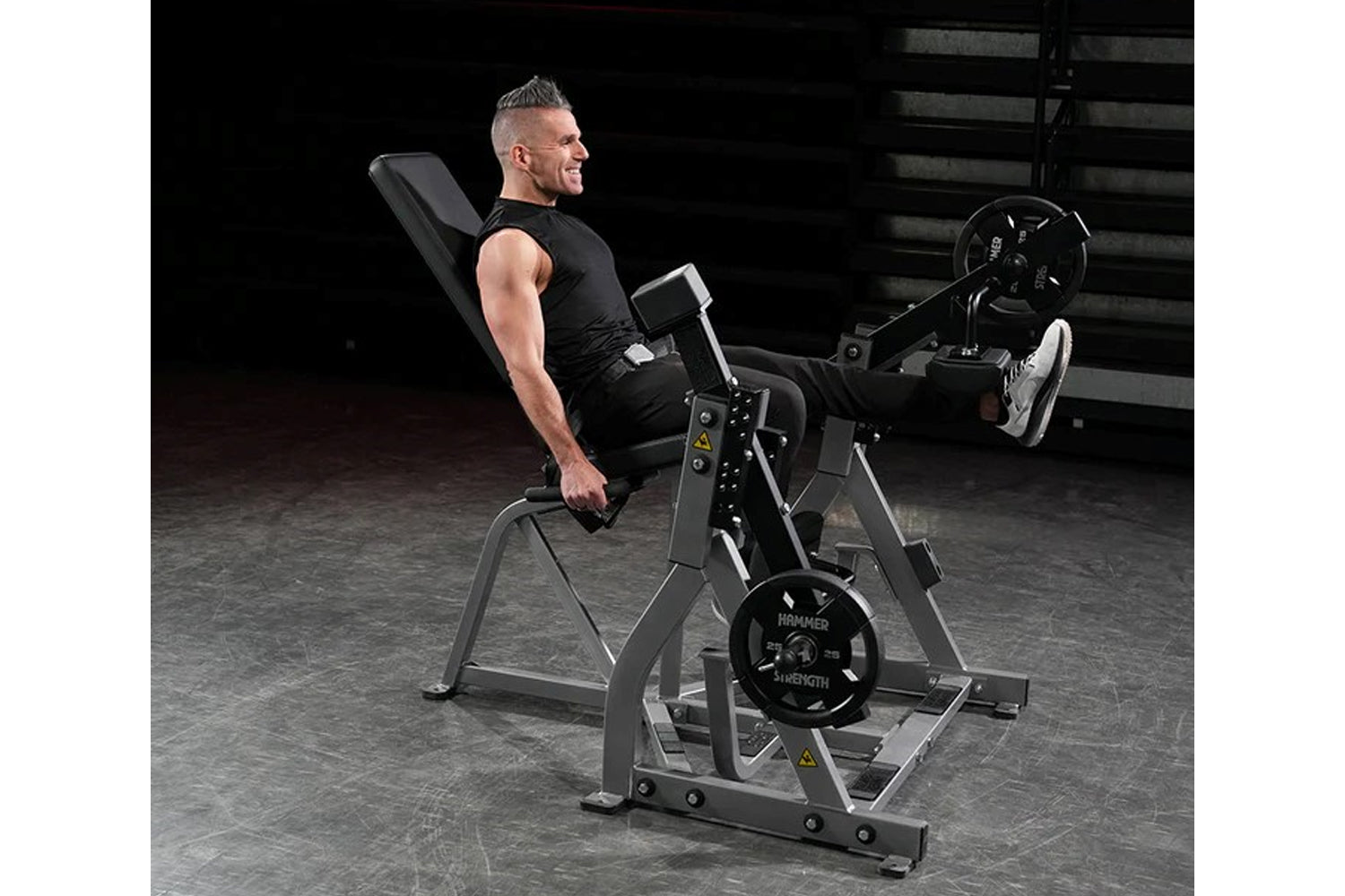Man in black sleeveless shirt using Hammer Strength plate-loaded leg extension machine with weighted plates in gym setting