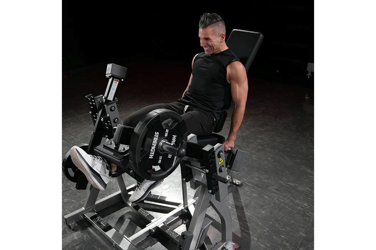 Man in black tank top using Hammer Strength plate-loaded leg extension machine with padded backrest and weight plates in gym 