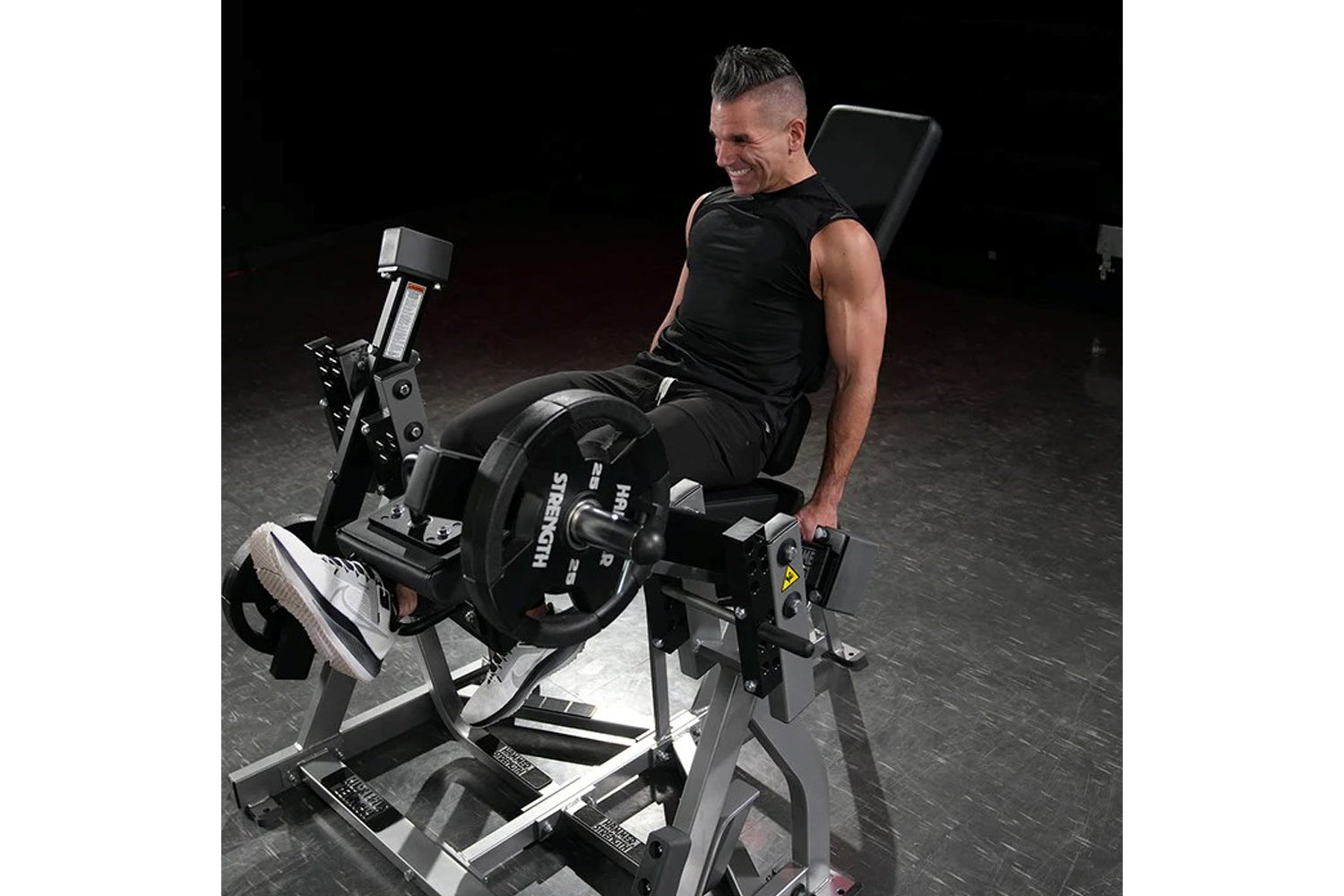 Man in black tank top using Hammer Strength plate-loaded leg extension machine with padded backrest and weight plates in gym 