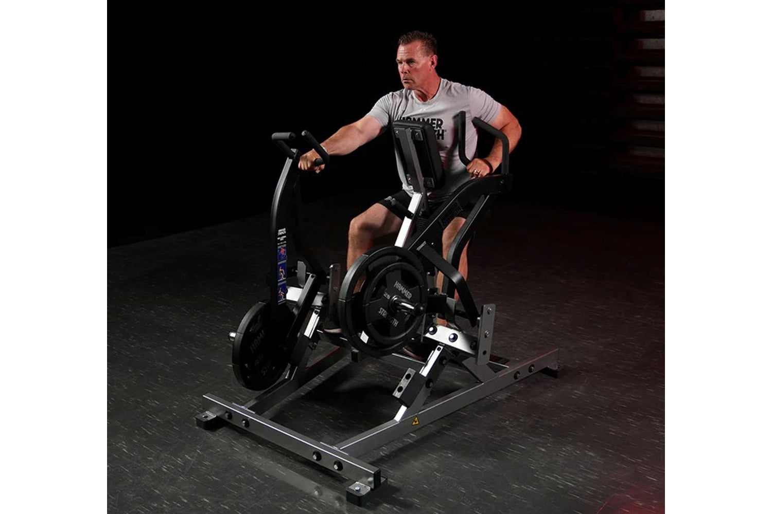 Man using black Hammer Strength plate-loaded iso lateral row machine, wearing gray t-shirt and black shorts on dark gym floor