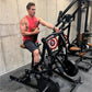Man in red tank top and navy shorts using black plate-loaded rowing machine in gym setting