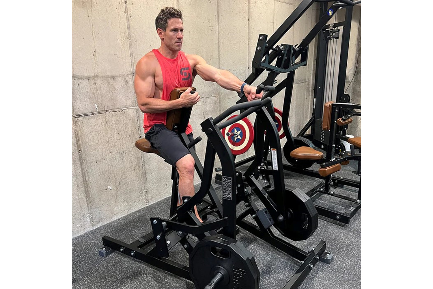 Man in red tank top and navy shorts using black plate-loaded rowing machine in gym setting