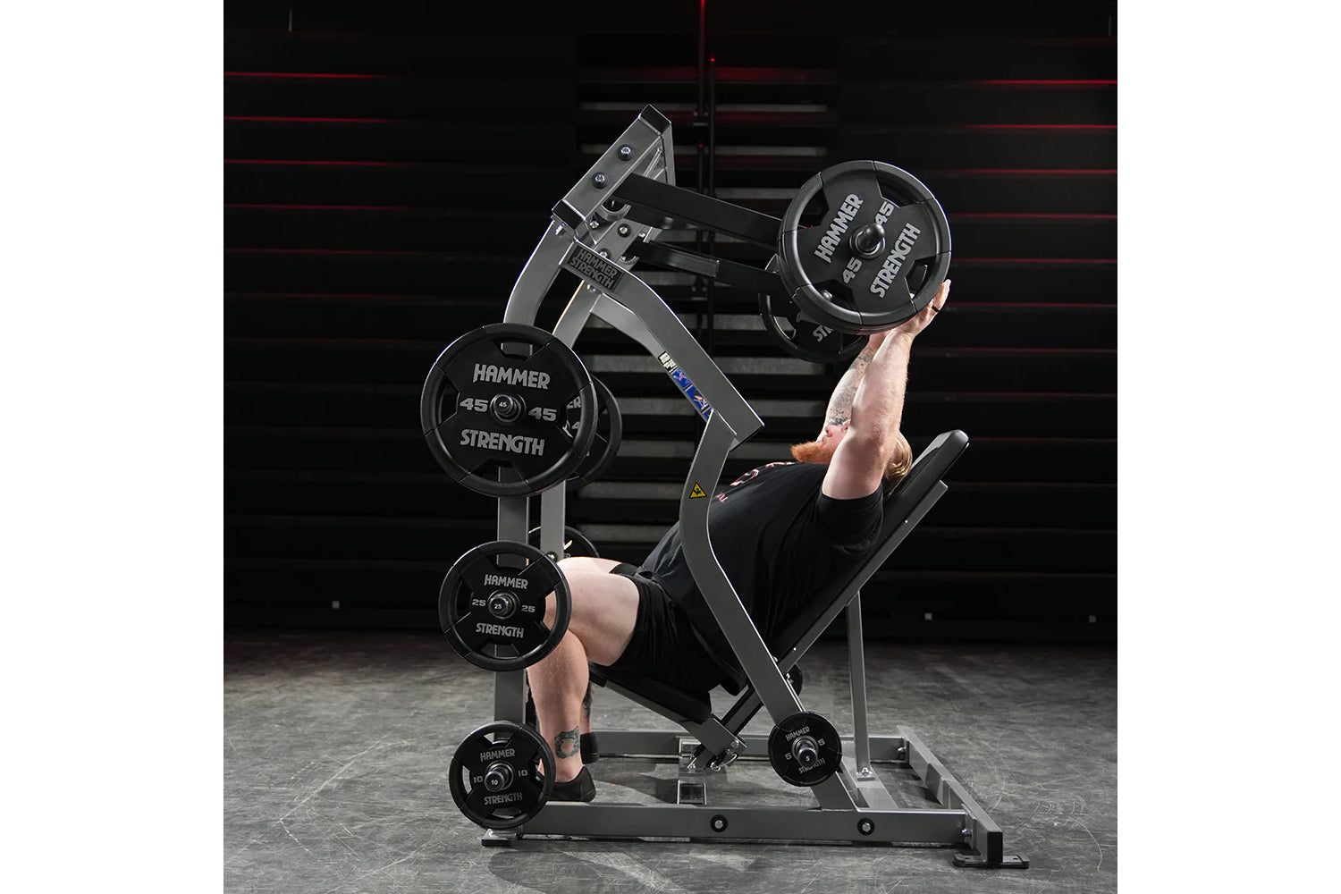 Man using Hammer Strength plate-loaded shoulder press machine in gym, lifting black weight plates overhead