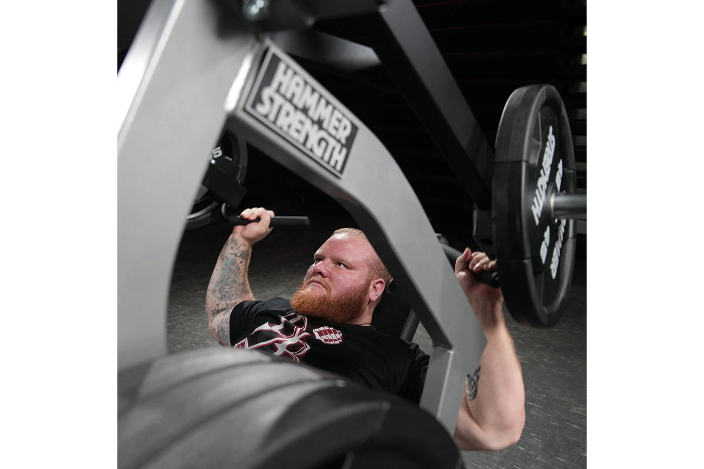Athlete with red beard and tattoos performing shoulder press on Hammer Strength plate-loaded machine with black weight plates