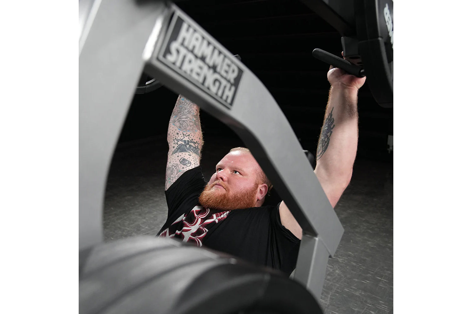 Man with red beard and chest tattoo using black Hammer Strength plate-loaded shoulder press machine in gym