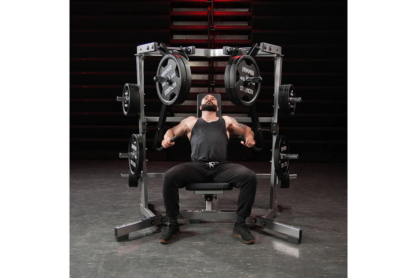 Bearded man in black tank top and pants performing chest press on Hammer Strength plate-loaded machine with loaded barbells i