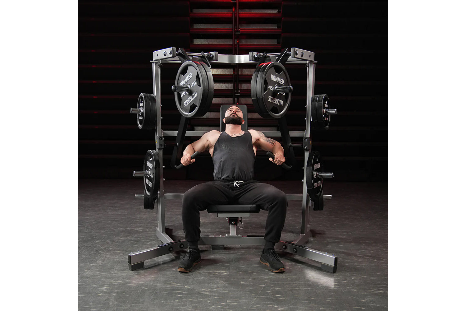 Bearded man in black tank top and pants performing chest press on Hammer Strength plate-loaded machine with loaded barbells i