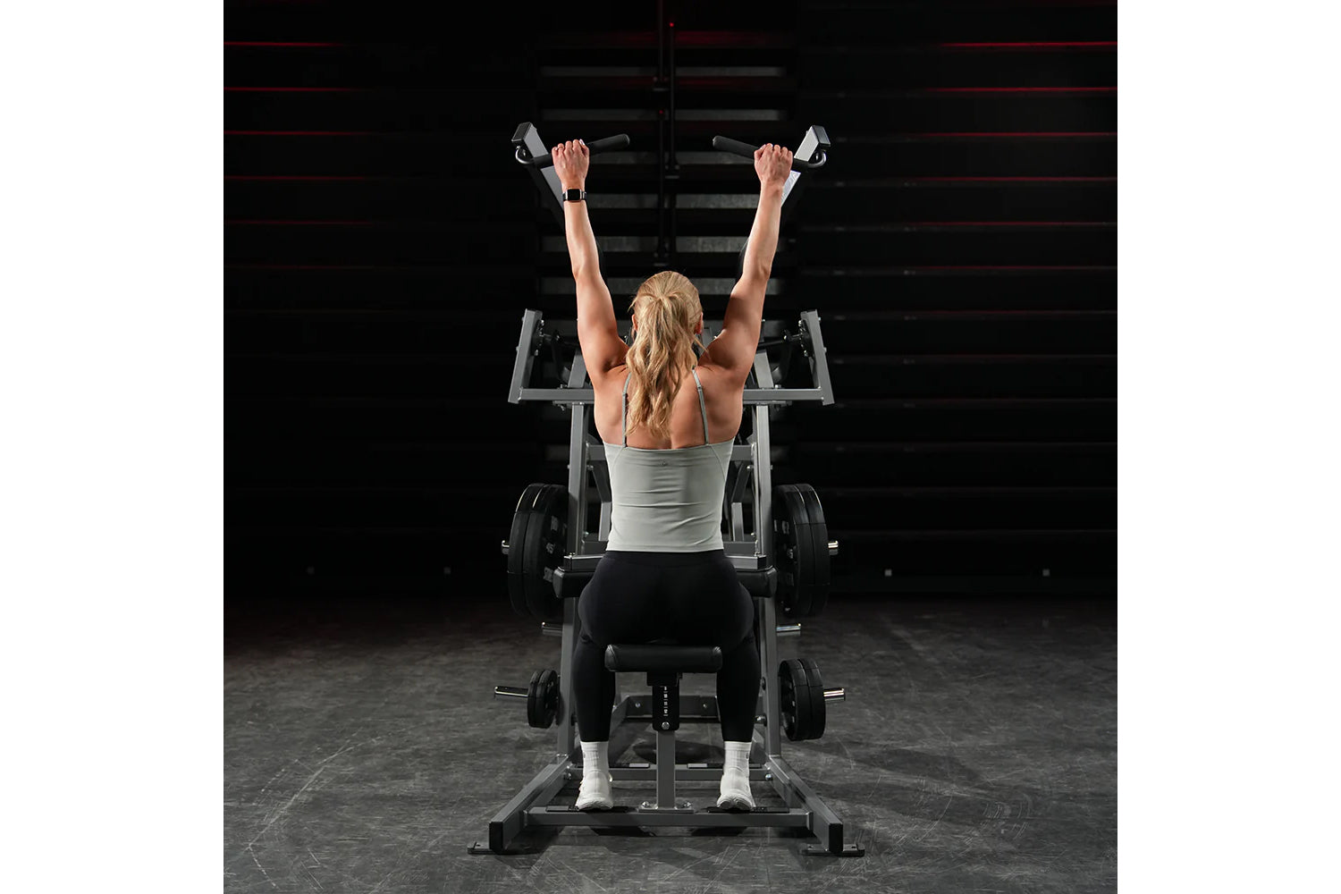Woman performing wide pulldown exercise on Hammer Strength plate-loaded machine, arms raised overhead gripping handles, weari
