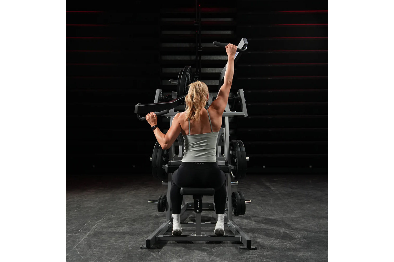 Woman performing wide pulldown exercise on Hammer Strength machine in gym setting, wearing gray tank top and black leggings