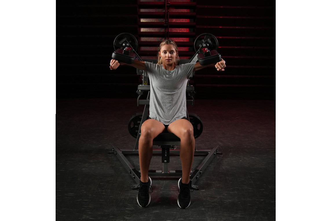 Woman performing lateral raise exercise on black Hammer Strength plate-loaded machine, holding dumbbells with arms extended, 