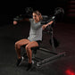Woman using black Hammer Strength plate-loaded lateral raise machine in gym setting, wearing gray t-shirt and black shorts