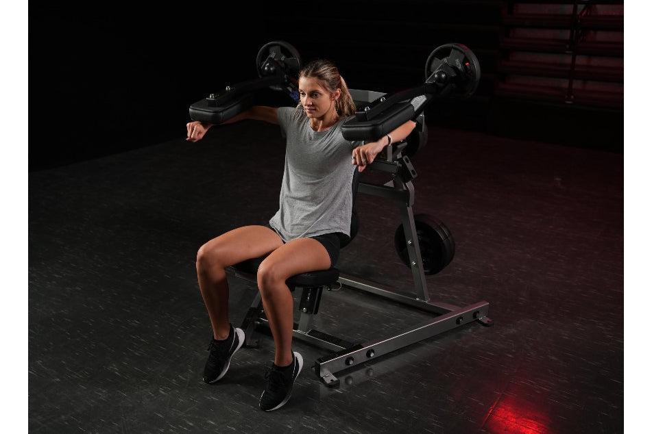 Woman using black Hammer Strength plate-loaded lateral raise machine in gym setting, wearing gray t-shirt and black shorts