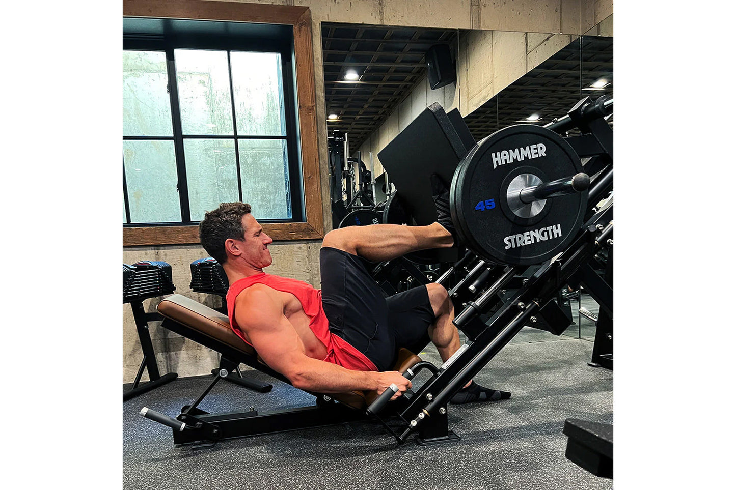 Man in red tank top using black Hammer Strength plate-loaded leg press machine with 45-pound weight plate in gym setting