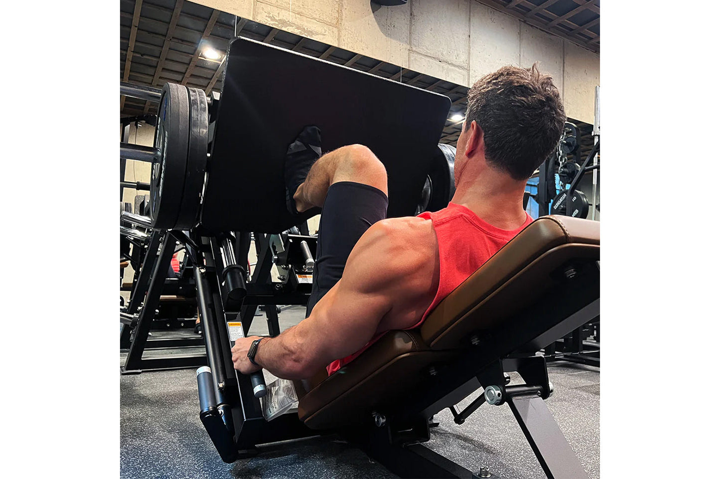 Man wearing red sleeveless tank top using black plate-loaded leg press machine in gym setting
