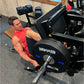 Man in red sleeveless shirt using black Hammer Strength plate-loaded leg press machine in gym setting
