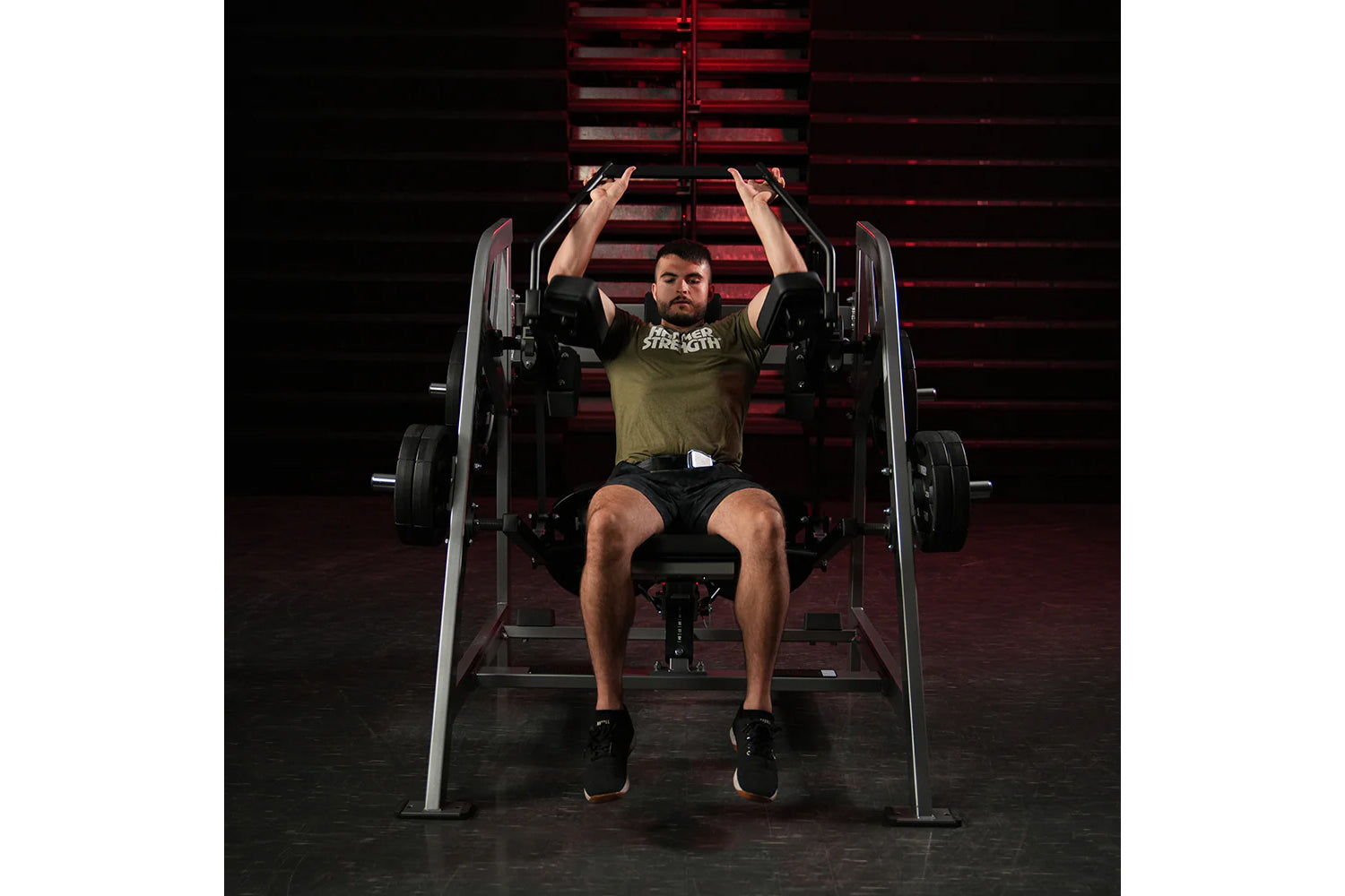 Man in olive t-shirt performing plate-loaded pullover exercise on Hammer Strength machine with red background