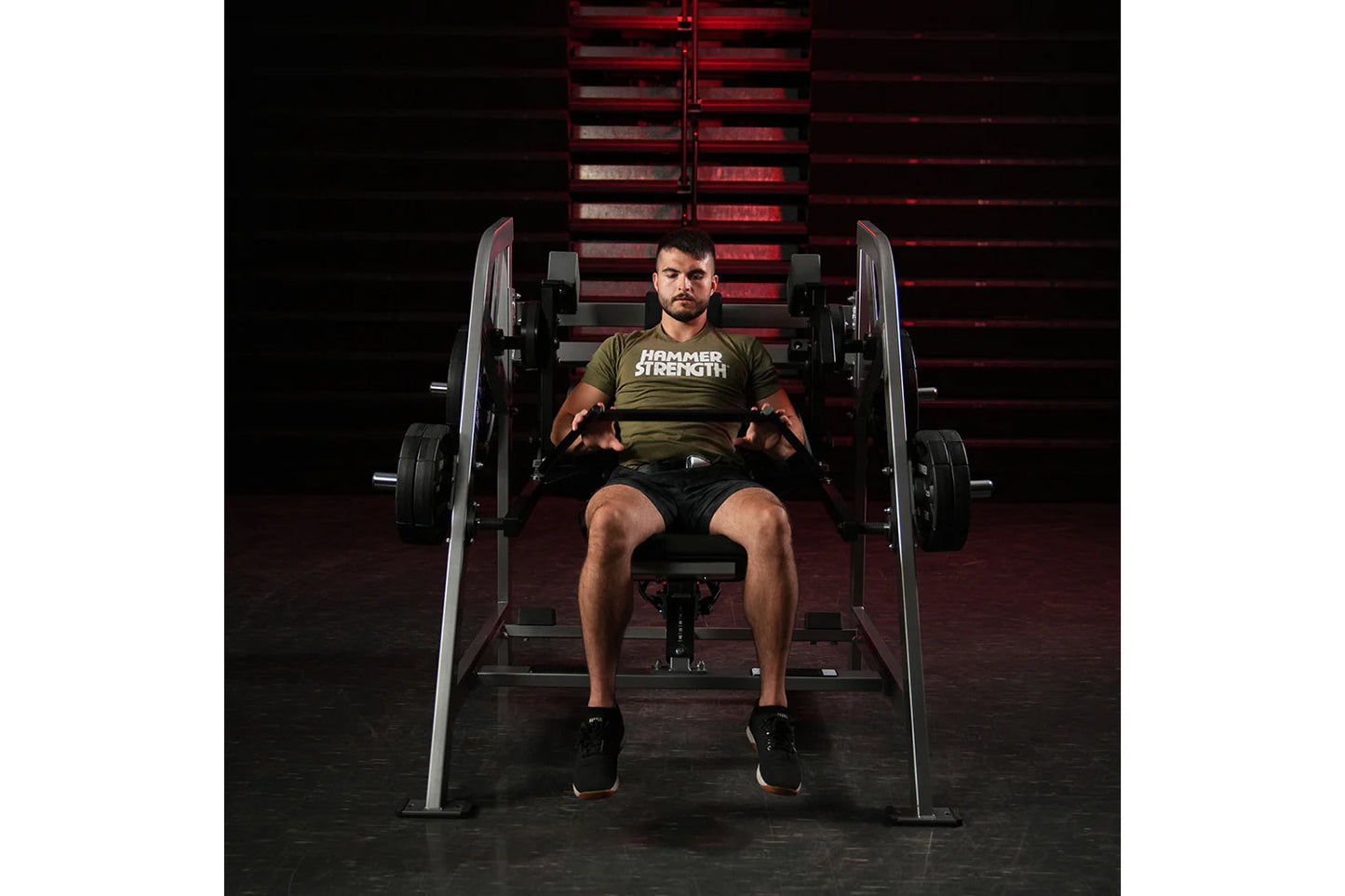 Man wearing olive Hammer Strength t-shirt using plate-loaded pullover machine in gym setting with red lighting