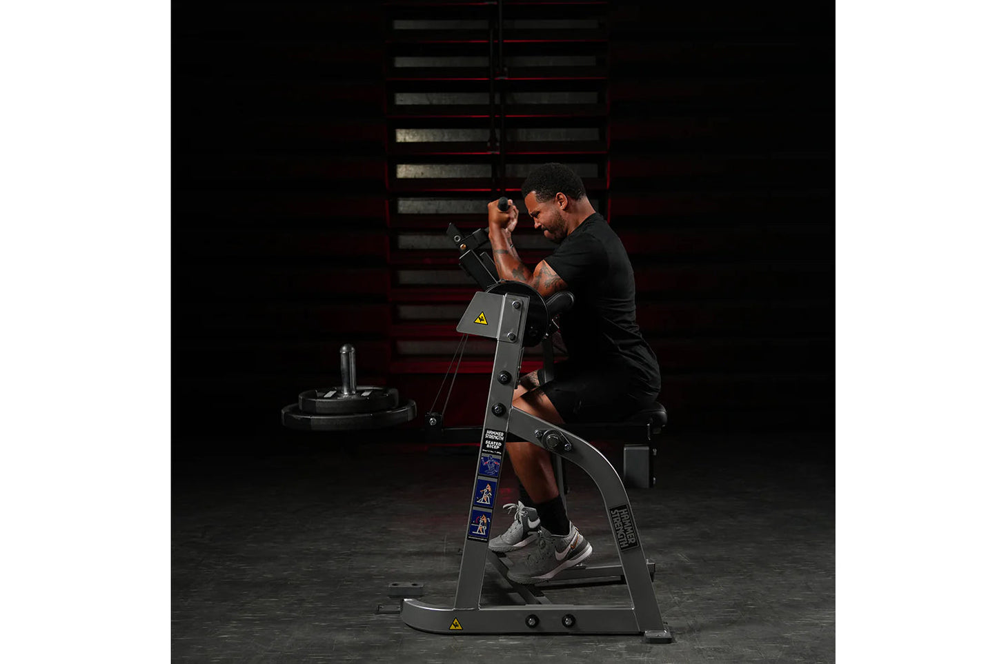 Man using black plate-loaded seated biceps machine in gym setting, demonstrating exercise form with weights loaded on machine