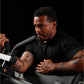 Man wearing black t-shirt demonstrating seated biceps machine with tattooed arms against dark background
