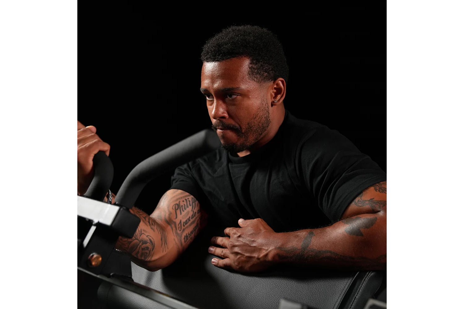 Man wearing black t-shirt demonstrating seated biceps machine with tattooed arms against dark background
