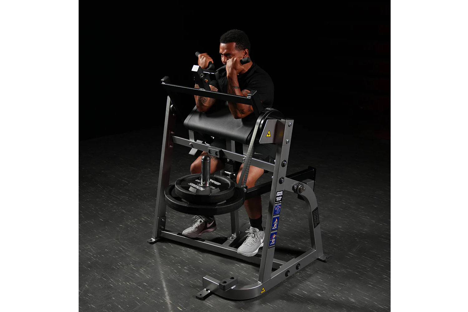 Man using black plate-loaded seated biceps machine with padded arm supports and pull handles against dark background