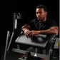 Man with tattoos using black Hammer Strength plate-loaded seated biceps machine with padded armrest against dark background