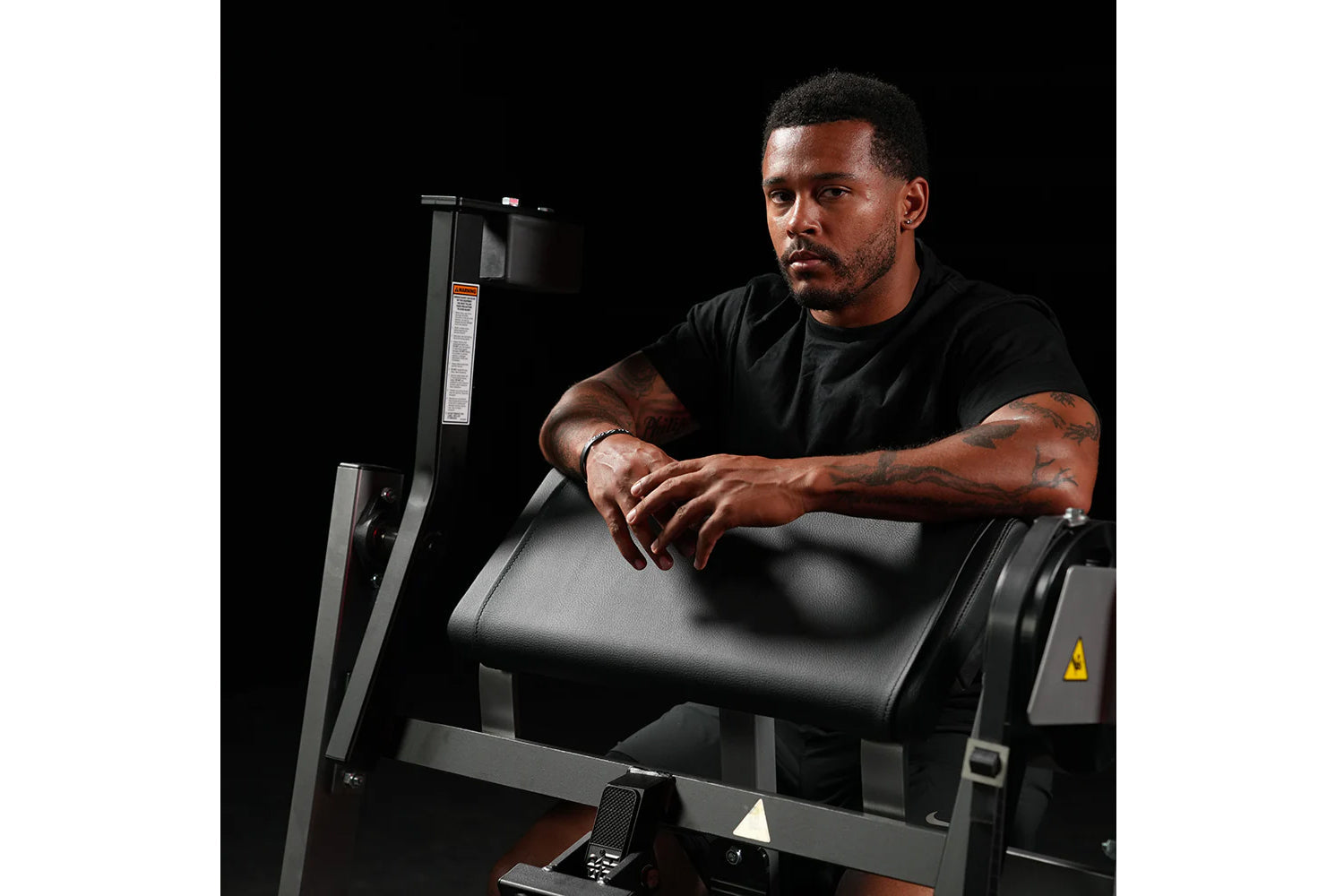 Man with tattoos using black Hammer Strength plate-loaded seated biceps machine with padded armrest against dark background