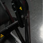 Black plate-loaded seated biceps machine with yellow caution labels and metal frame, detail view of mechanical arm and handle