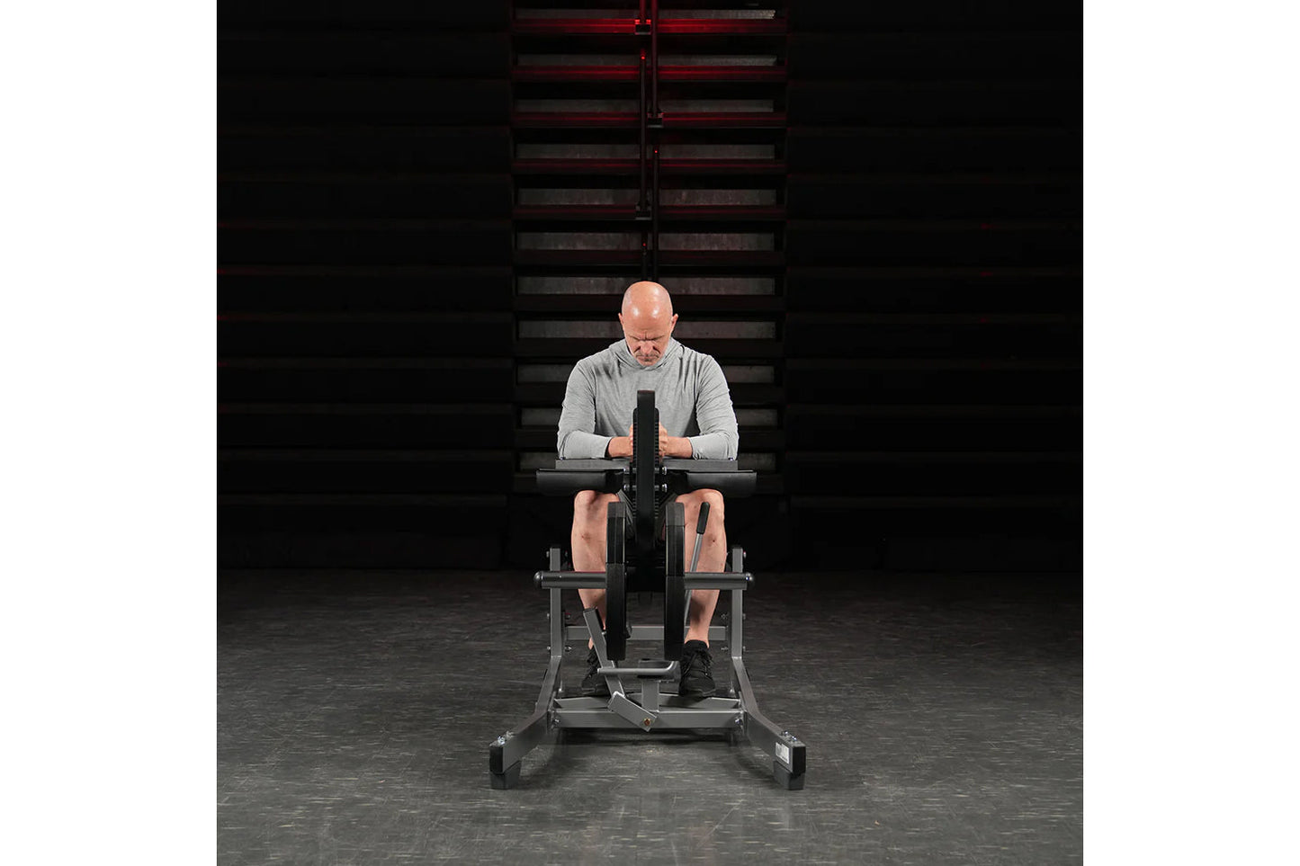 Man seated on Hammer Strength plate-loaded calf raise machine wearing gray long-sleeve shirt and black shorts in gym setting