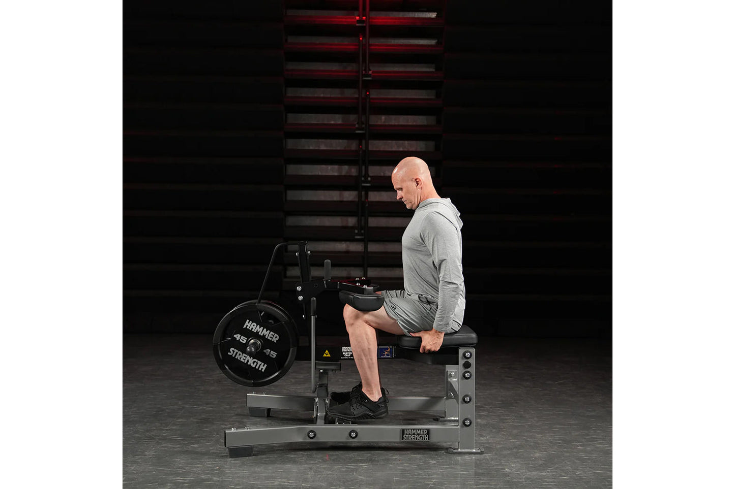 Man in gray long-sleeve shirt and black shorts using Hammer Strength plate-loaded seated calf raise machine in gym setting