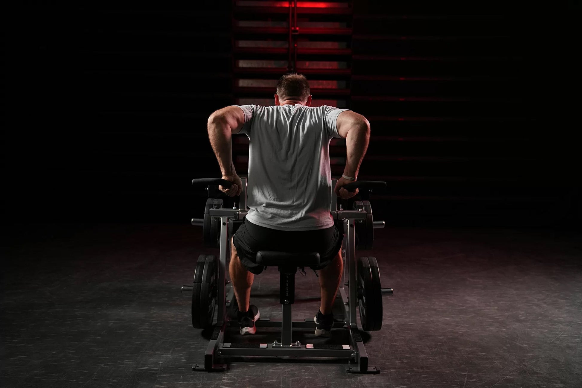 Man wearing gray sleeveless tank top using plate-loaded seated dip machine in gym setting with red lighting