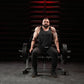 Man wearing black sleeveless tank top and black pants seated on Hammer Strength plate-loaded shrug machine with dumbbells, in