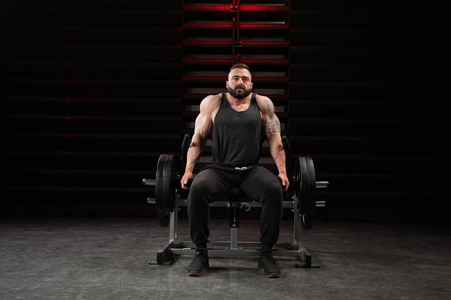 Man wearing black sleeveless tank top and black pants seated on Hammer Strength plate-loaded shrug machine with dumbbells, in